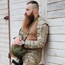 Man wearing MFH multi-terrain BDU combat shirt holding helmet, leaning against wooden wall in tactical gear