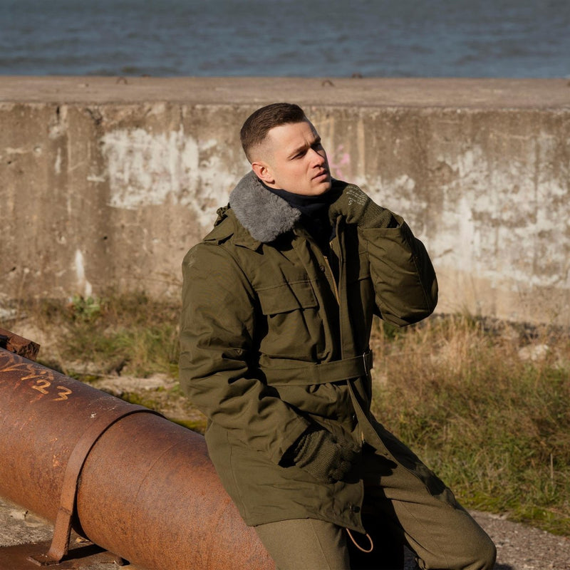 Man in a green jacket sitting on a rusted pipe by a concrete wall and water.